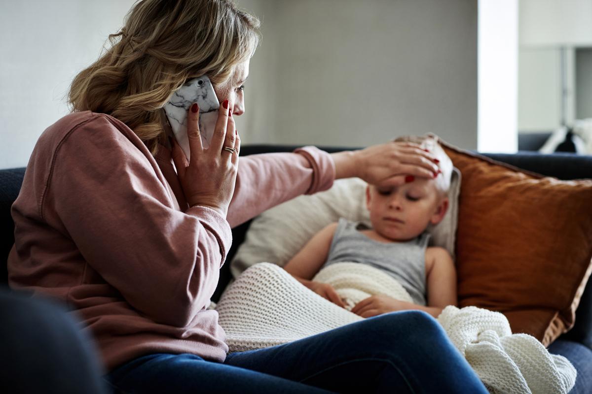 Woman on phone with doctor while checking son's forehead temperature with her hand.